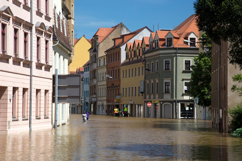 A flooded outdoor area near a modern building with large glass windows and exterior lighting, featuring a significant accumulation of water covering the concrete surface and part of the sidewalk. The 