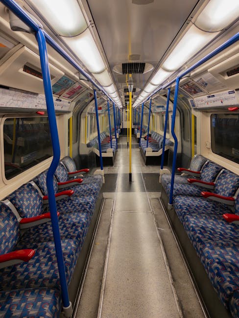 The interior of an empty train carriage displaying a long central aisle flanked by rows of upholstered seats with blue and red patterned fabric. The carriage features yellow and blue handrails, silver metal poles, and overhead lighting illuminating the space. The windowed sides are clean, with dark reflections visible on the glass. The floor appears well-maintained, free of dust or debris, highlighting a sanitized environment typical of professional cleaning by Cleaners Ilford, prioritizing hygiene and surface cleanliness in public transportation settings.