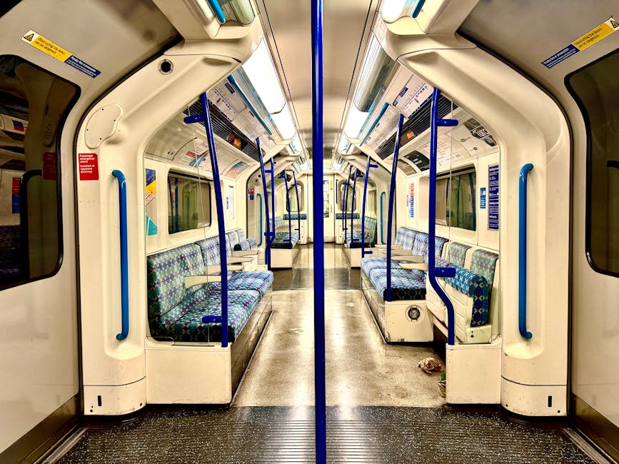 The interior of an empty train carriage featuring rows of upholstered seats with patterned blue and green fabric, set against white plastic and metal walls. Blue handrails run along the top and sides, with vertical support bars positioned near the doors and seats. The floor appears clean and slightly textured, with some minor debris visible near the door threshold. Large windows with clear glass line both sides of the carriage, allowing natural light to illuminate the space. Overhead shelves and informational signage are visible above the seats. The lighting is bright and even, highlighting the cleanliness and orderly arrangement of the seating area, typical of a professional cleaning or sanitisation process by Cleaners Ilford, as advertised on the page for Gants Hill station carpet and upholstery cleaning options in Ilford.