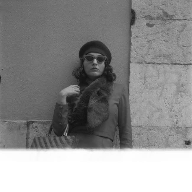 A woman dressed in a stylish coat with a fur collar, wearing dark sunglasses and a beret, stands with a confident expression against a textured stone wall and smooth plastered surface. She holds a handbag with striped detailing over her shoulder, and the lighting highlights the neat folds of her coat and the polished surface of her accessories. The background suggests an urban outdoor setting with subtle lighting emphasizing the cleanliness and well-maintained appearance of the environment. This image illustrates a sense of modern design and personal presentation, relevant to residential or commercial cleaning needs in the context of hygiene and maintenance standards, as emphasized on the page about avoiding hidden charges in deep cleaning quotes at cleaners-ilford.com.