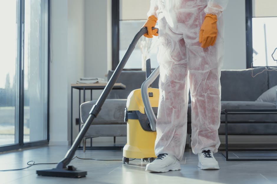 A professional cleaner dressed in white disposable coveralls, orange gloves, and white sneakers is operating a yellow and grey vacuum cleaner on a dark tiled floor in a modern living room. The room features a grey fabric sofa, a black side table with a phone or remote on top, and large glass sliding doors allowing natural light to illuminate the space. The floor appears clean and dry, with the vacuum actively being used to remove dust and debris, emphasizing surface cleaning and sanitisation typical of deep cleaning processes involved in residential cleaning with attention to hygiene standards, as offered by Cleaners Ilford. The scene focuses on maintaining a spotless environment through thorough surface cleaning, highlighting the importance of professional deep cleaning services to avoid hidden charges, as explained on the page