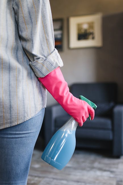 Close-up of a person wearing a light blue striped shirt and pink rubber gloves, holding a blue spray bottle with a green trigger in their right hand, ready for surface cleaning. The background shows a modern living room with a dark grey sofa, and framed pictures on a grey wall, illuminated by natural light. This scene depicts thorough cleaning practices typically performed during domestic cleaning or end of tenancy cleaning, emphasizing hygiene and surface sanitation. The focus is on the individual's hand and cleaning tools, highlighting the prepare-for-departure cleaning process that may include disinfecting furniture and surfaces, consistent with detailed deep cleaning services offered by Cleaners Ilford, as referenced on the page about end of tenancy cleaning prices and hidden fees.