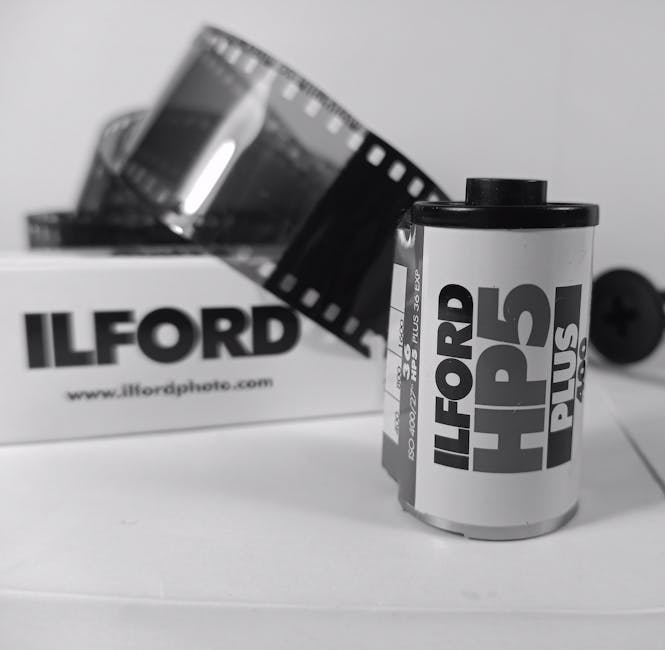 A close-up of a 35mm film roll labeled 'Ilford HP5 Plus', standing upright on a white surface in front of a box with 'Ilford' branding and a partially unwound strip of film. The background includes a blurred black camera lens and a screw, highlighting a professional photography setup. The scene is well-lit, emphasizing the glossy texture of the film roll and the smooth surface of the box, with minimal dust or smudges visible on the film material. This image illustrates high-quality surface and product cleaning by Cleaners Ilford, ensuring the preservation and hygiene of photographic equipment and supplies, consistent with their focus on detailed cleaning services for both residential and commercial environments.