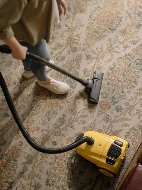A person using a yellow vacuum cleaner with a black hose attachment to deep clean a patterned area rug in a room. The rug features floral motifs in shades of beige, green, and red. The individual, wearing beige shoes and dark jeans, holds the vacuum upright on the carpeted surface. The room is well-lit, and nearby furniture is partially visible. The scene demonstrates surface cleaning and maintenance, aligning with professional services offered by Cleaners Ilford, as referenced on the page for same-day carpet cleaning in Ilford.