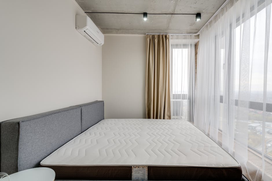 A modern bedroom featuring a neatly made bed with a gray upholstered headboard and a clean, white mattress. The room has large floor-to-ceiling windows covered with sheer white curtains and beige drap