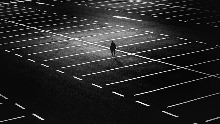 A person standing alone in a nearly empty parking lot illuminated by a single overhead light, with neatly painted white lines marking the parking spaces on the dark asphalt surface. The scene emphasizes cleanliness and surface maintenance, highlighting the smoothness and brightness of the painted lines against the clean, dark asphalt. The lighting creates stark contrasts, and no vehicles are present, giving an impression of hygiene and tidiness in an outdoor environment. This setting showcases the importance of surface cleaning and upkeep, aligning with domestic and commercial cleaning practices promoted by Cleaners Ilford for maintaining hygienic outdoor surfaces. The overall scene suggests careful surface cleaning and sanitisation to ensure cleanliness.
