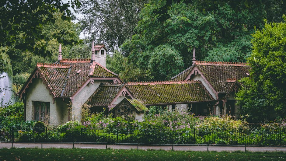 A charming, historic house with a steep, moss-covered red-tiled roof and decorative woodwork embellishing the gables, situated among lush green trees and foliage. The house features light-colored stucco walls with dark-framed windows, surrounded by a well-maintained garden filled with flowering plants and shrubbery, enclosed by a black metal fence. Natural daylight illuminates the scene, highlighting the clean, pristine condition of the house exterior, indicative of thorough surface cleaning and maintenance by professional services such as Cleaners Ilford. The setting exudes a peaceful, well-kept atmosphere suitable for domestic cleaning and garden upkeep within the Valetines Park area in Ilford.